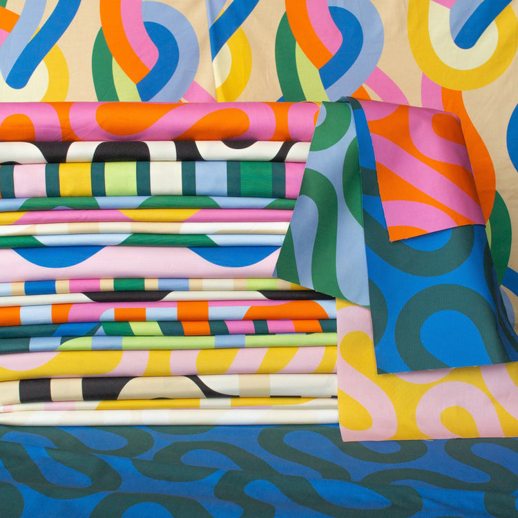 A stacked and draped display of the Groove collection by Emily Van Hoff, featuring bold, curvy, interwoven patterns in vibrant colors like blue, green, yellow, pink, and orange